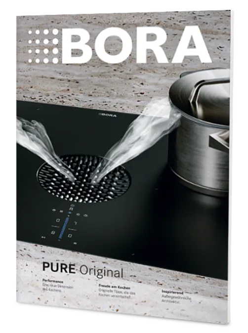 Titelbild des BORA Magazins 2025: Moderne Küche mit orangefarbener Insel, balancierend auf einem Felsen. Schwarze Backöfen und Steinwand.