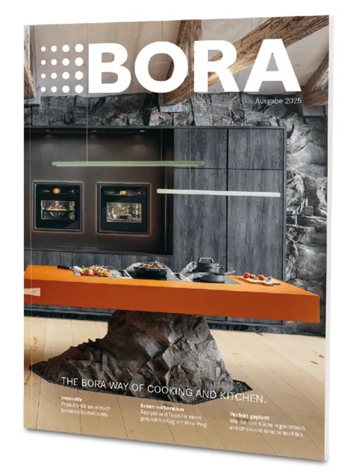 Titelbild des BORA Magazins 2025: Moderne Küche mit orangefarbener Insel, balancierend auf einem Felsen. Schwarze Backöfen und Steinwand.