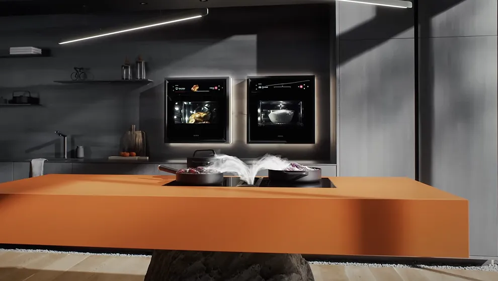 Moderne Küche in Grau-Orange. Eine leuchtend orangefarbene Kücheninsel mit schwarzem Induktionskochfeld. Zwei Töpfe dampfen, Backöfen im Hintergrund.