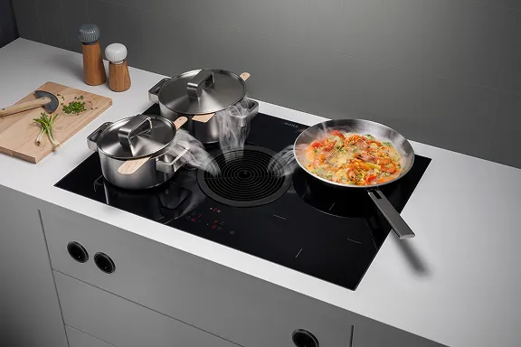 Detail: Schwarzes BORA Induktionskochfeld mit rundem Abzug in weißer Platte. Paella-ähnliches Gericht kocht. Dampf wird in der Mitte abgesaugt.